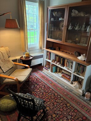 A cozy room with a chair, a bookshelf, a radiator, and a window with curtains.