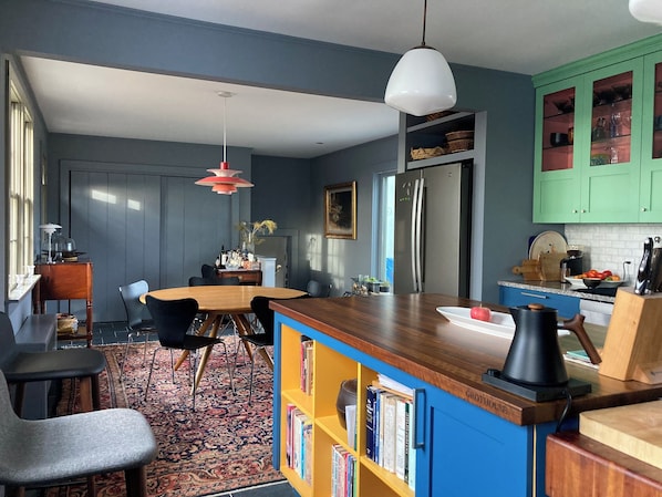 A modern kitchen with a central island, a dining area with chairs, a bookshelf, and a refrigerator.
