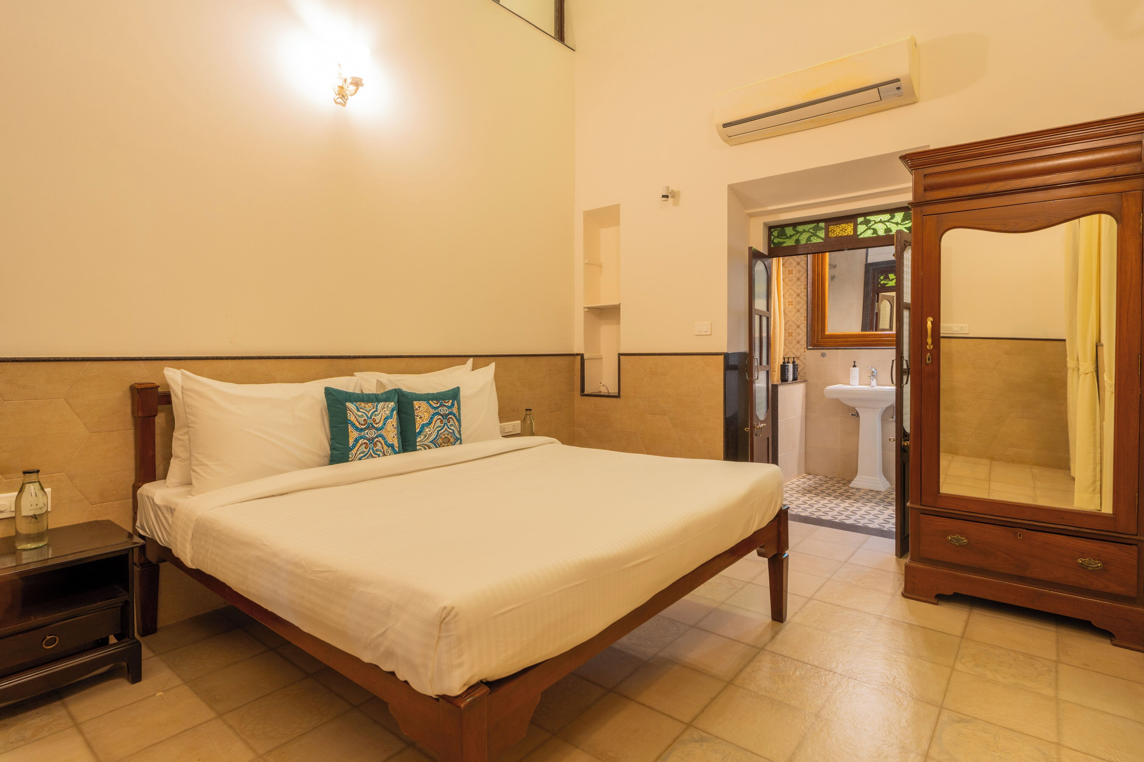 Habitació d'hotel amb un llit gran, una tauleta de nit de fusta, un armari, un bany amb rentapans i mirall, i una unitat de aire condicionat a la paret.