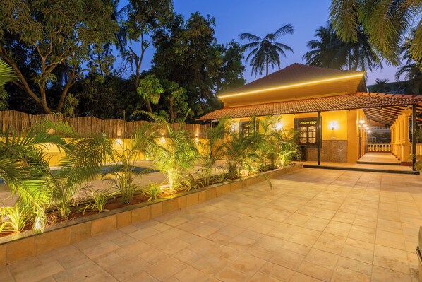 A well-lit courtyard with a tiled floor, surrounded by plants and palm trees, leading to a building with a tiled roof and a wooden fence.