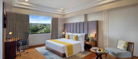Habitación de hotel con una cama grande, un escritorio, una silla, una mesita con fruta y vistas a la ciudad.