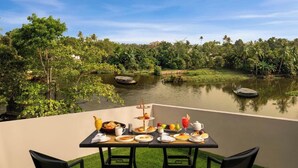A dining table with breakfast items and chairs, overlooking a calm lake with trees and a clear sky. - Olivia Alleppey (Alappuzha)