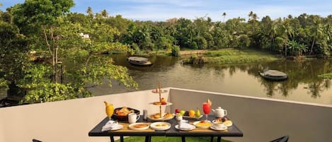 A dining table with breakfast items and chairs, overlooking a calm lake with trees and a clear sky.