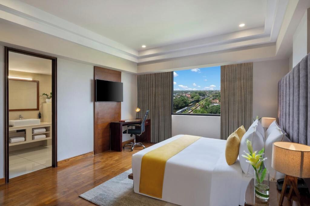 A hotel room with a bed, a desk, a TV, a bathroom, and a view of a cityscape.
