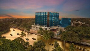 A modern hotel with a glass facade, situated by a river with boats. - Olivia Alleppey (Alappuzha)