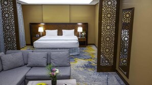 Une chambre d’hôtel avec un grand lit, un canapé, une table basse avec des fruits et un panneau décoratif au mur.