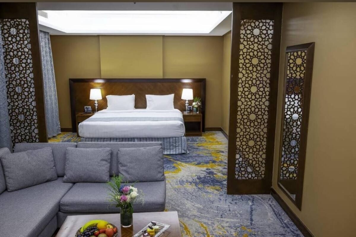 A hotel room with a large bed, a sofa, a coffee table with fruit, and a decorative wall panel.