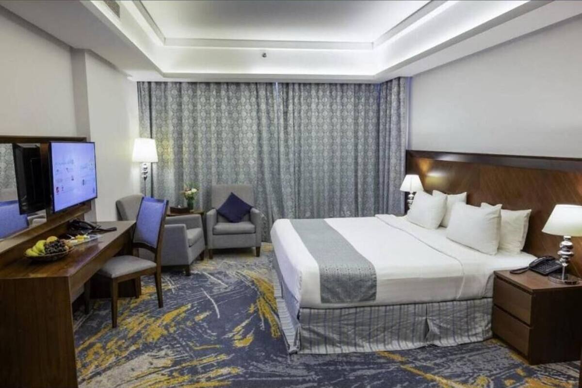 A hotel room with a bed, desk, chair, TV, lamps, and a patterned carpet.