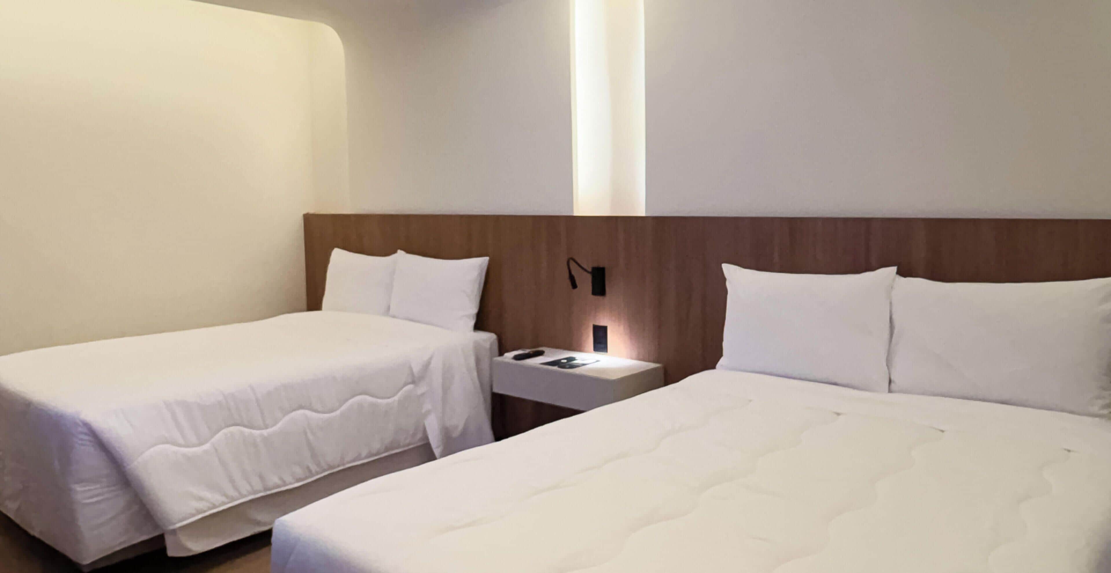A hotel room with two single beds, a nightstand with a lamp, and a wall-mounted light fixture.