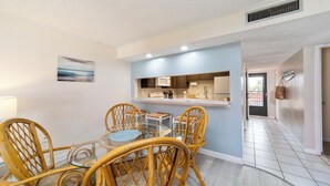 A dining area with a glass table and wicker chairs, a kitchen in the background, and a hallway leading to another room. - Pelican Inlet C222: steps to pool, tennis; boat ramp and storage available (Saint Augustine)