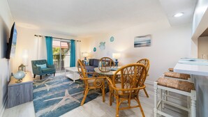A living room with a sofa, a dining table with chairs, a TV mounted on the wall, and a window with curtains. - Pelican Inlet C222: steps to pool, tennis; boat ramp and storage available (Saint Augustine)