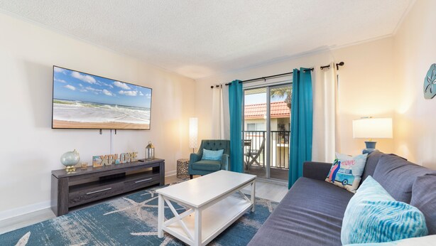 A living room with a flat-screen TV, a white coffee table, a grey sofa, teal curtains, and a view of the outside. - Pelican Inlet C222: steps to pool, tennis; boat ramp and storage available (Saint Augustine)