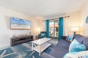 Un salon doté d’un téléviseur à écran plat, d’une table basse blanche, d’un canapé gris, de rideaux turquoise et offrant une vue sur l’extérieur.