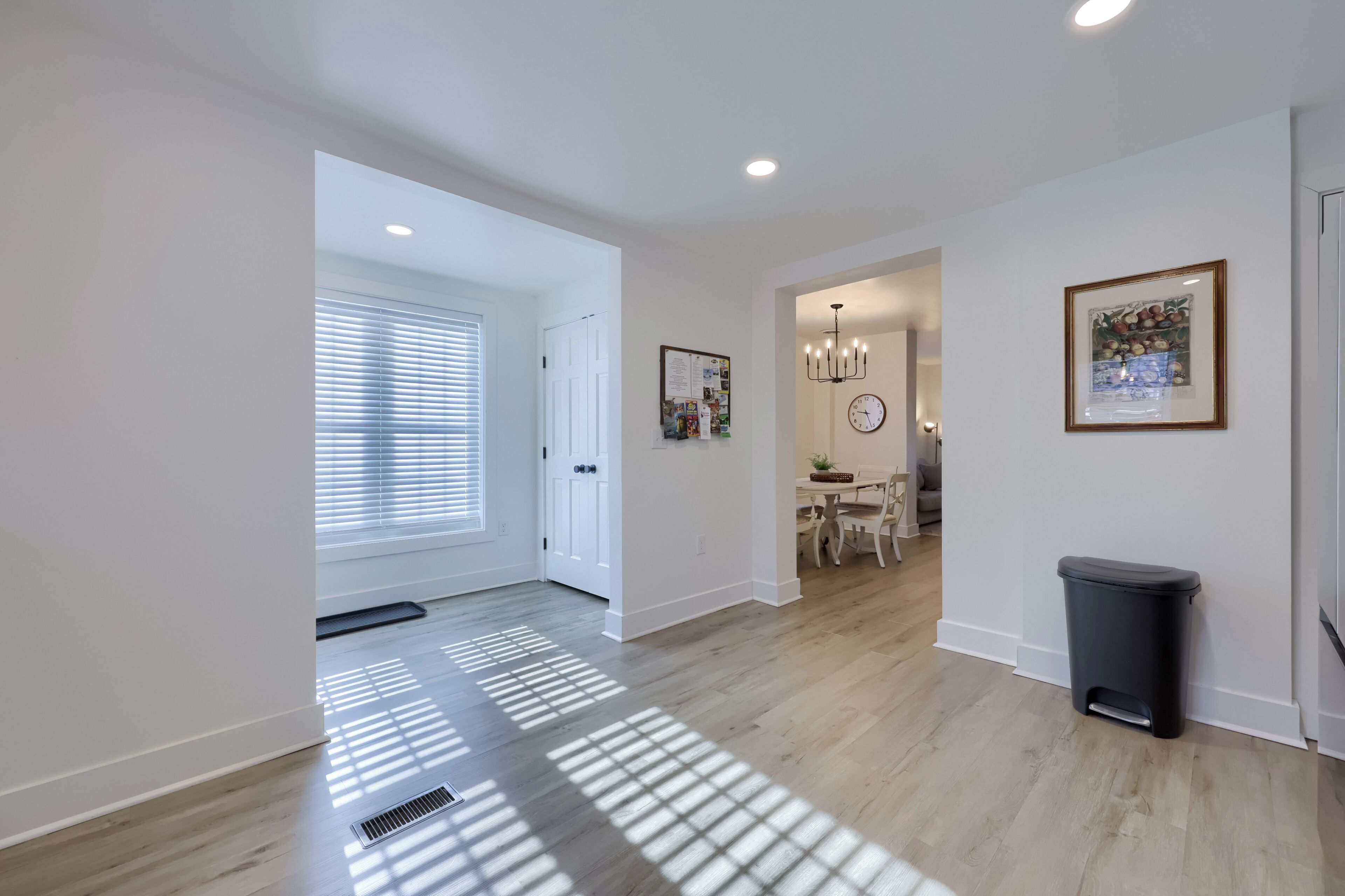 A spacious hallway with a dining area in the background, a trash bin, and a wall-mounted clock.