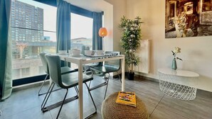 A modern living room with a dining area, large windows, a potted plant, and a magazine on a round ottoman. - Suite in Antwerp Near Meir Shops (Antwerp)