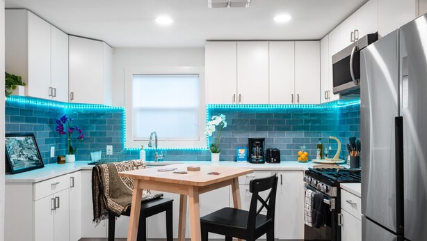 A modern kitchen with white cabinets, a stainless steel refrigerator, a wooden table, and blue LED lights along the backsplash. - East Austin Bungalow w BBQ Firepit Fun Yard (Austin)