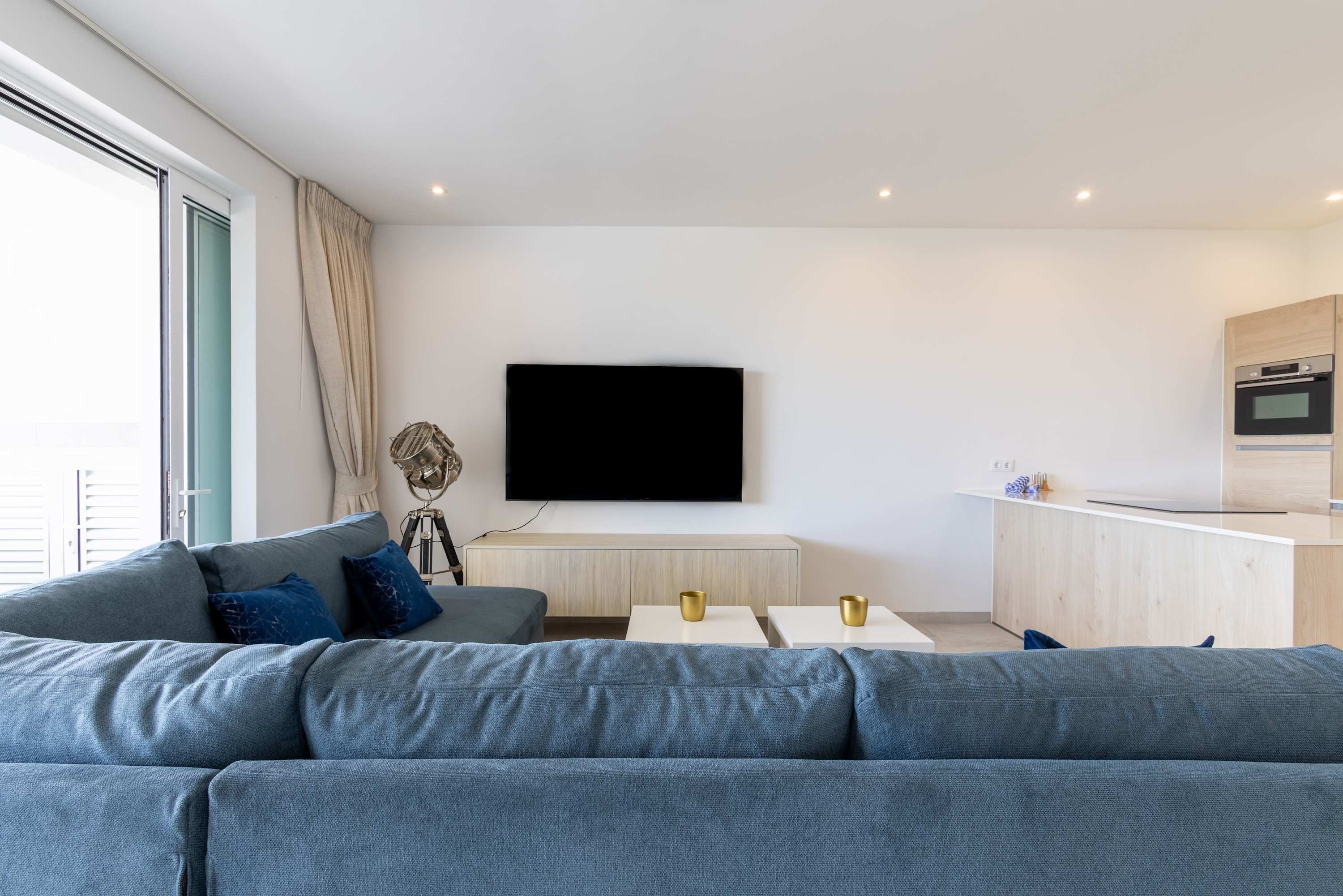 Una sala de estar moderna con un sofá gris, un televisor de pantalla plana montado en la pared y una zona de cocina con armarios de madera y una encimera.