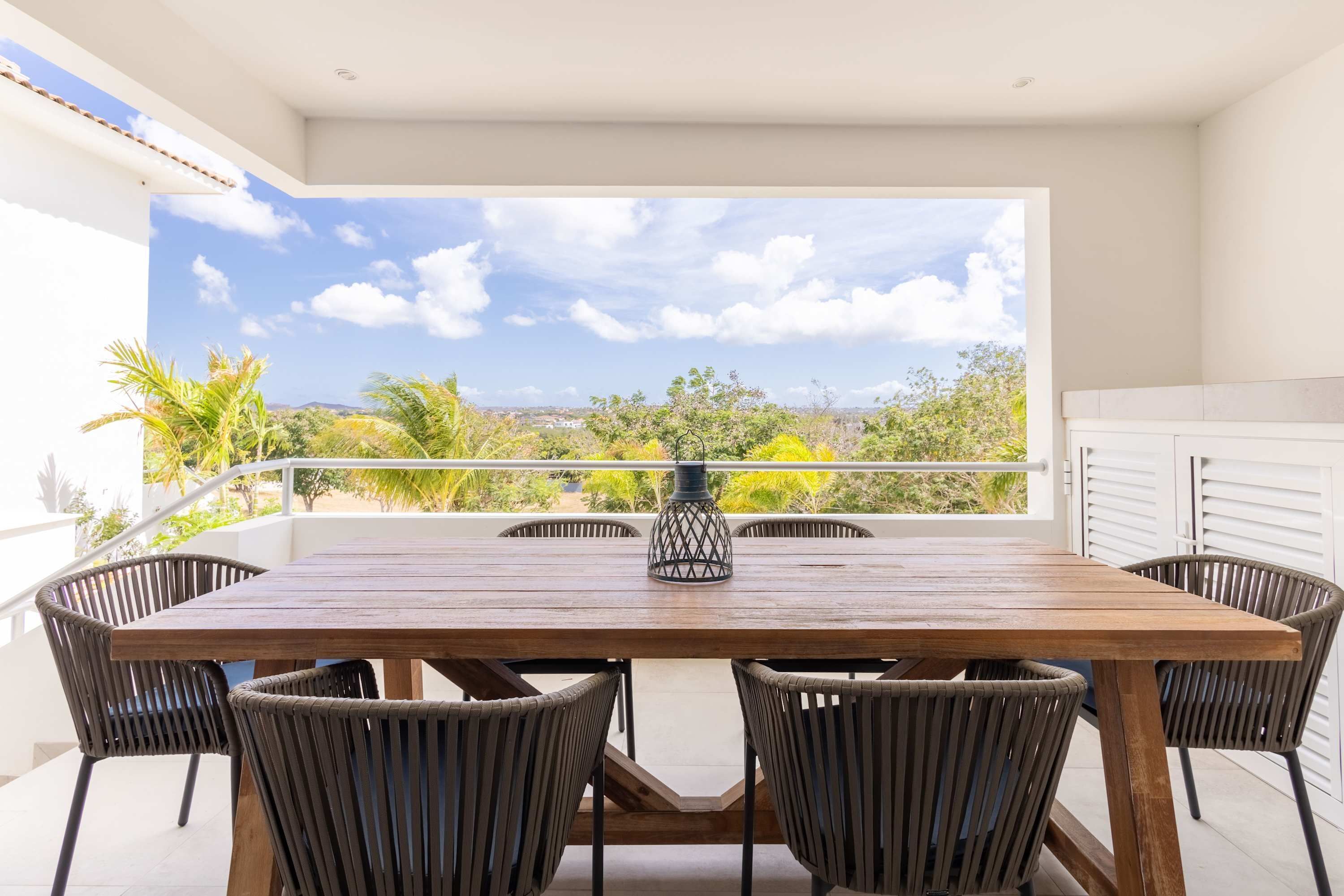 A wooden table with six chairs on a balcony with a view of greenery and a clear sky.