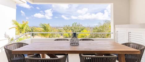 A wooden table with six chairs on a balcony with a view of greenery and a clear sky.