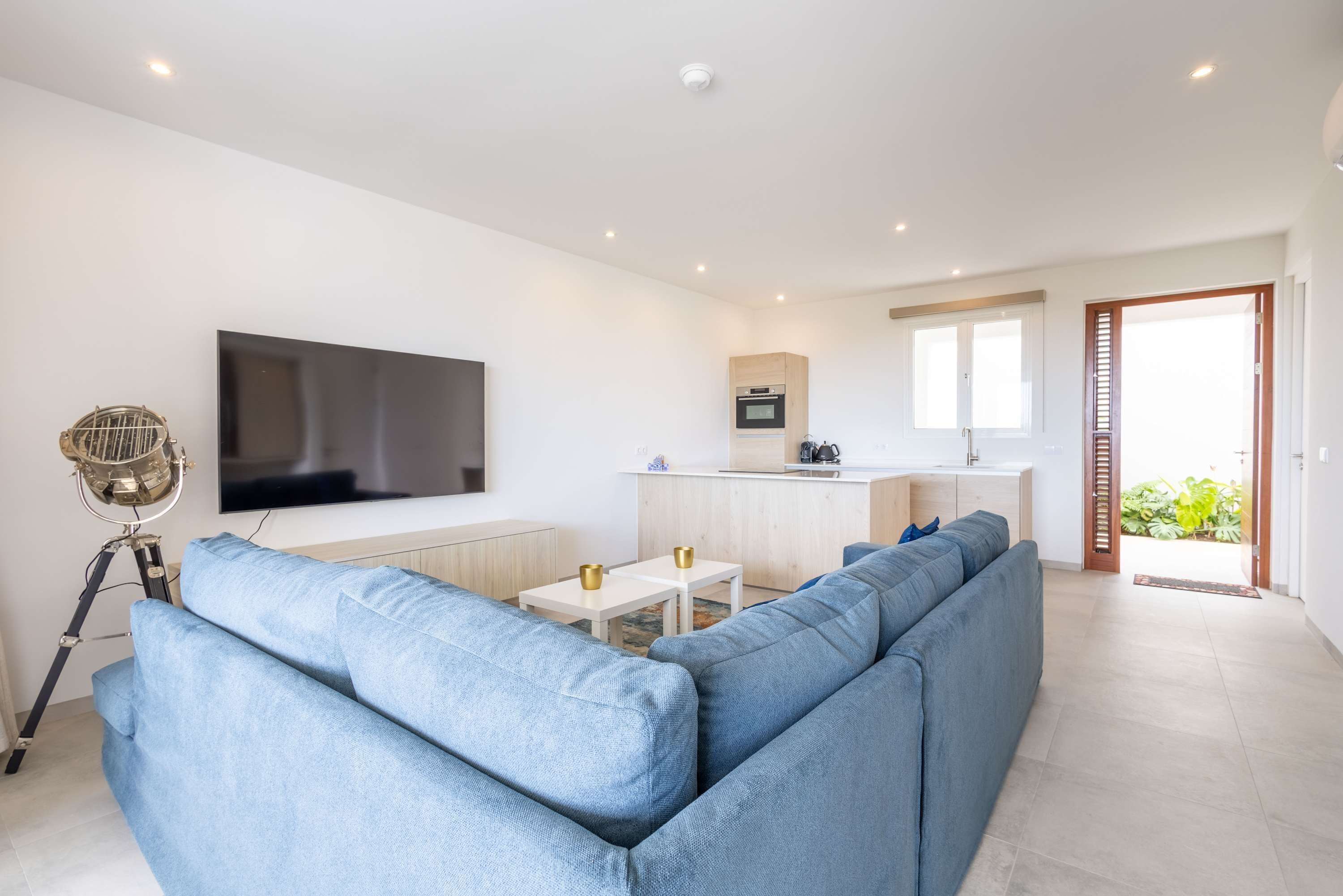 A modern living room with a sectional sofa, a flat-screen TV, a small kitchen area, and a sliding glass door leading to a balcony.