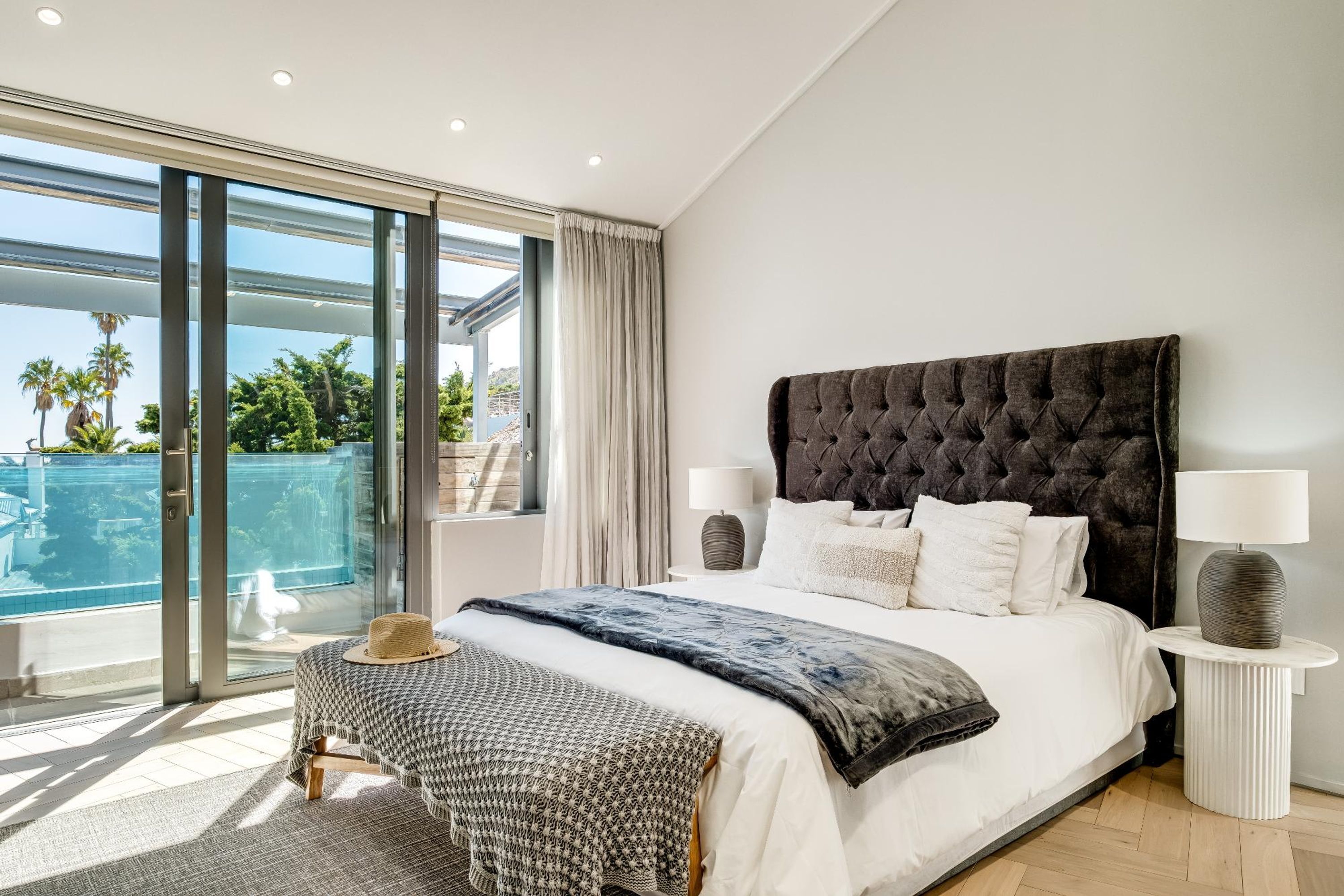 Een slaapkamer met een groot bed, twee nachtkastjes met lampen, uitzicht op een zwembad en een balkon.
