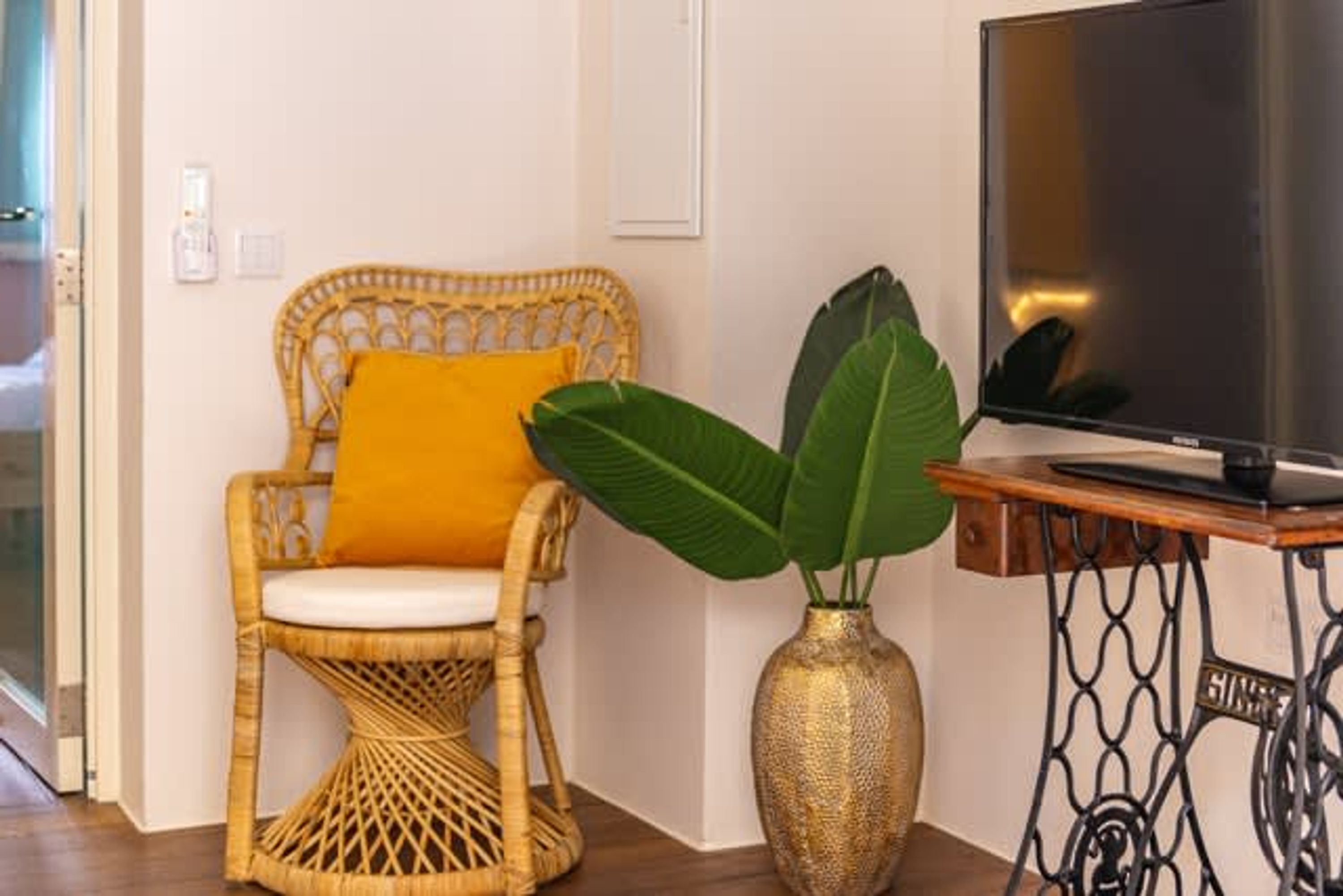 Uma cadeira de vime com almofada, uma grande planta verde em vaso dourado e uma televisão sobre uma mesa de madeira.