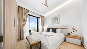 A modern bedroom with a large bed, a bedside table, a bench, and a view of the city. - Free Housekeeping Visits During Your Stay - Stayshort - Relaxing 1 Bedroom in JVC That Sleeps 2 (Dubai)