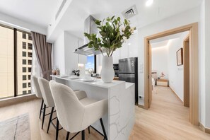 A modern kitchen with a white island, bar stools, a large vase with greenery, and a view of a cityscape through large windows. - Free Housekeeping Visits During Your Stay - Stayshort - Relaxing 1 Bedroom in JVC That Sleeps 2 (Dubai)