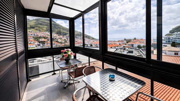 A balcony with a table and chairs, a flower vase, and a view of a town with red-roofed buildings and hills. - Apartament Brigitte in Machico (Machico)