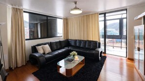 A modern living room with a black leather sectional sofa, a glass coffee table, and a large window offering a city view. - Apartament Brigitte in Machico (Machico)