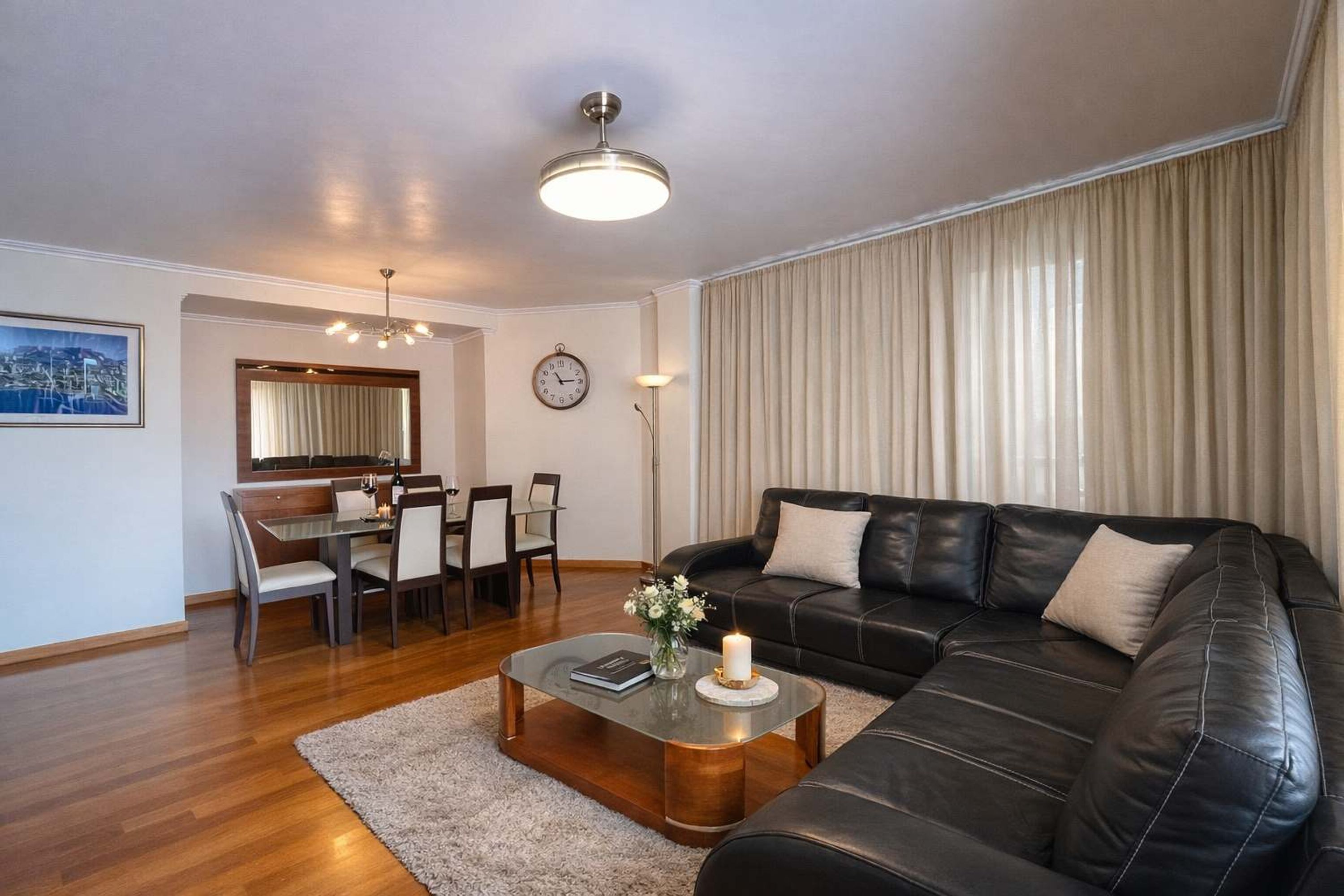 A modern living room with a black leather sofa, a glass coffee table, a dining area with a wooden table and chairs, a large window with curtains, and a cityscape painting on the wall.