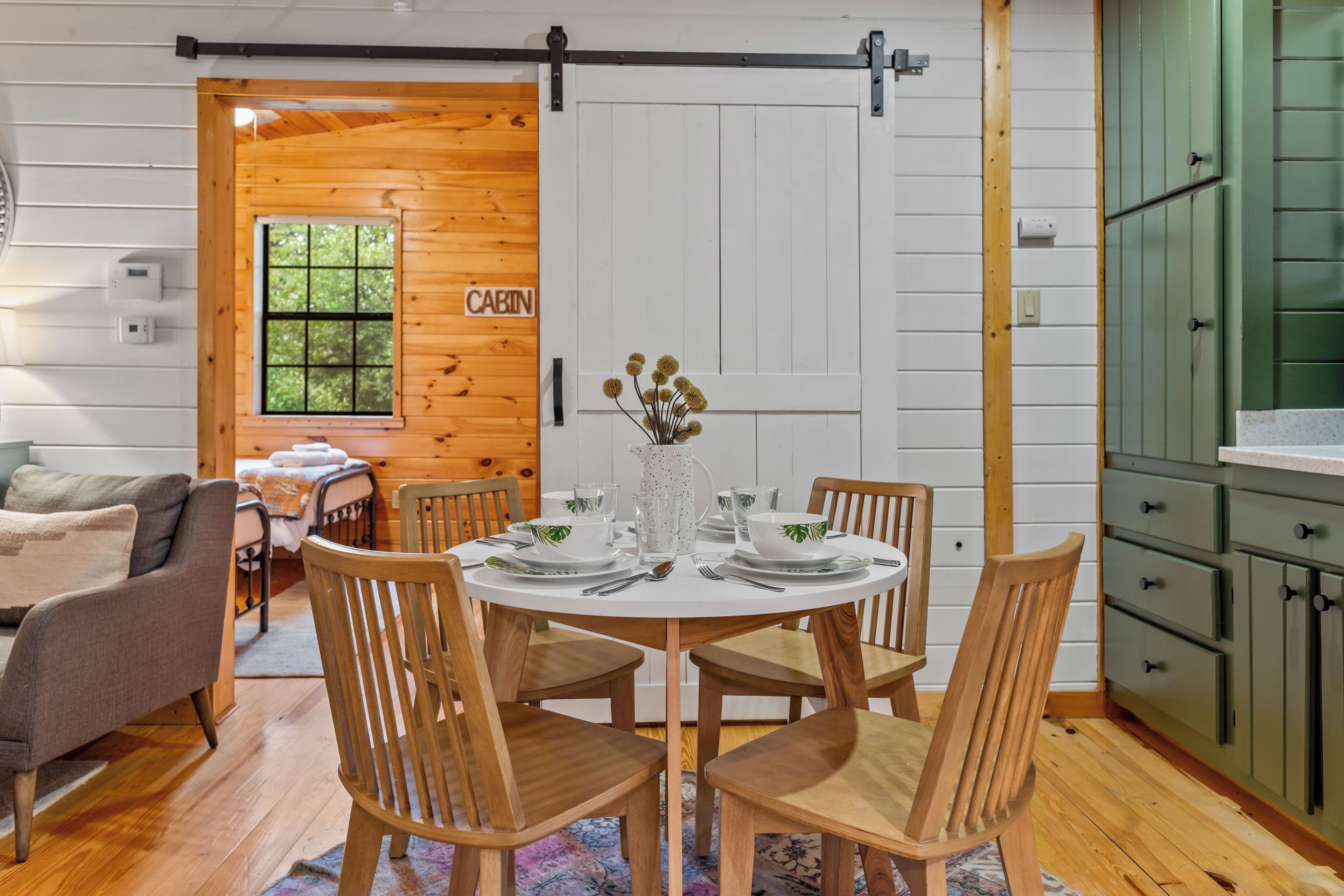 A cabin interior with a dining table set for four, wooden chairs, a kitchen area with green cabinets, and a 'CABIN' sign on the door.