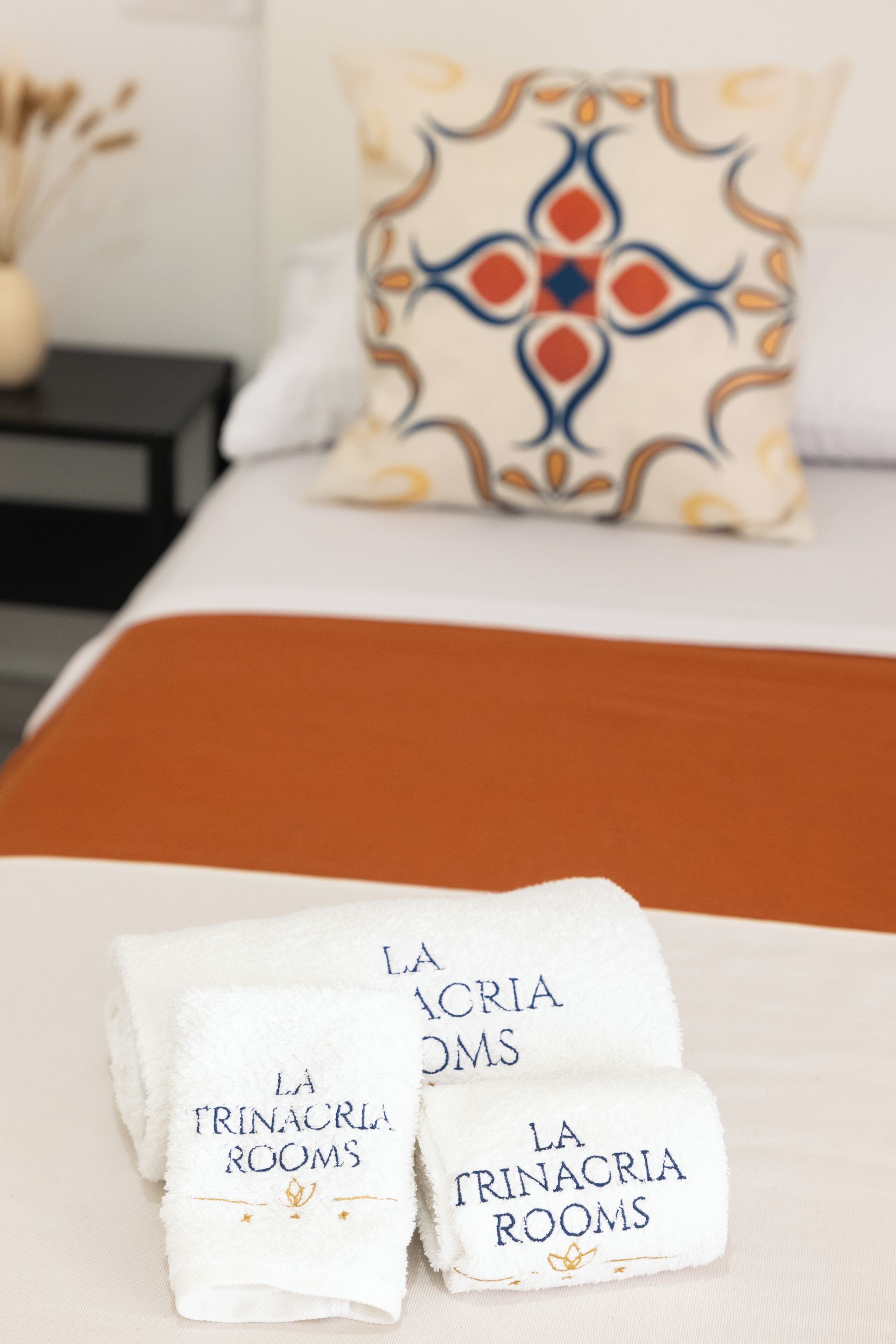 A neatly made bed with a white and orange bedspread, two folded towels with text, and a decorative pillow in the background.
