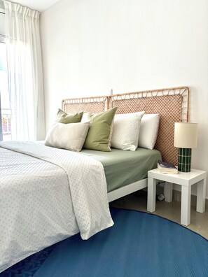 Una cama bien hecha con una colcha verde, almohadas blancas y un cabecero blanco. Hay una mesita de noche blanca con una lámpara y una alfombra azul en el suelo.
