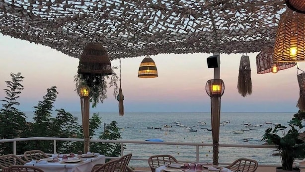 An outdoor dining area with wicker furniture and tables set for a meal, overlooking a body of water with boats. - Private Cove Salions (Tossa de Mar)