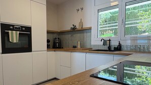 A modern kitchen with white cabinetry, a built-in oven, a coffee maker, and a window with a view of greenery. - Private Cove Salions (Tossa de Mar)