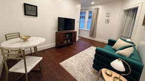 A living room with a TV, a dining table with chairs, a sofa, and a window with curtains. - Renovated 2BR - Workspace - Near UC Med & Cincy (Saint Bernard)