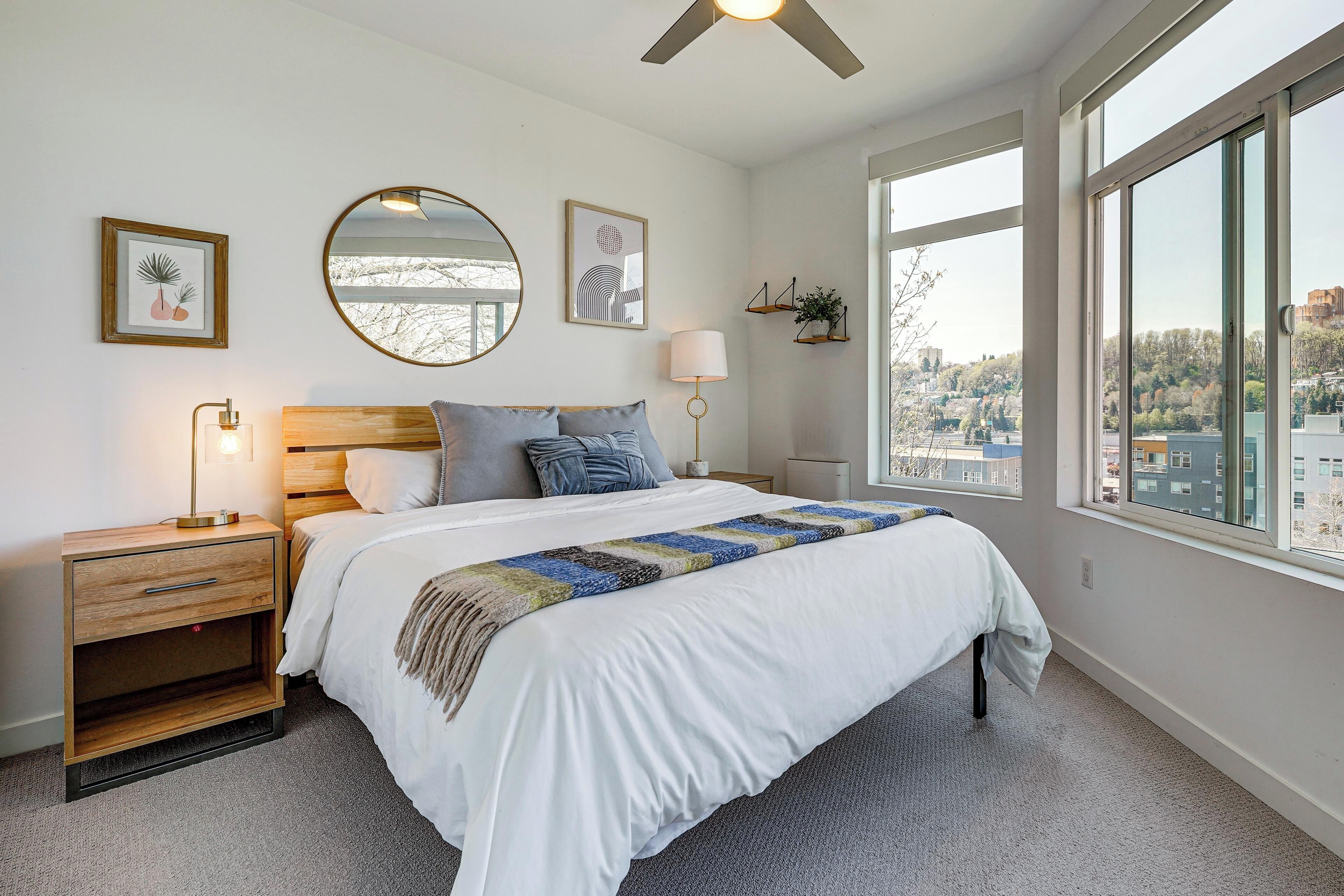 A bedroom with a bed, bedside tables, a mirror, wall art, and a window with a city view.