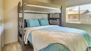 Una cama litera con colcha blanca y azul turquesa, cabecera de madera y una ventana con persianas.