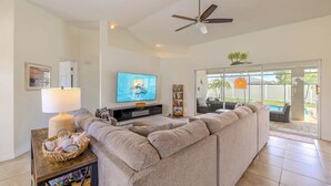 A living room with a large sofa, a TV, a ceiling fan, a lamp, a bookshelf, and a view of the outdoors. - Bring your dogs and enjoy the charm of this home! 3 be- 2ba in SE Cape Coral (Cape Coral)