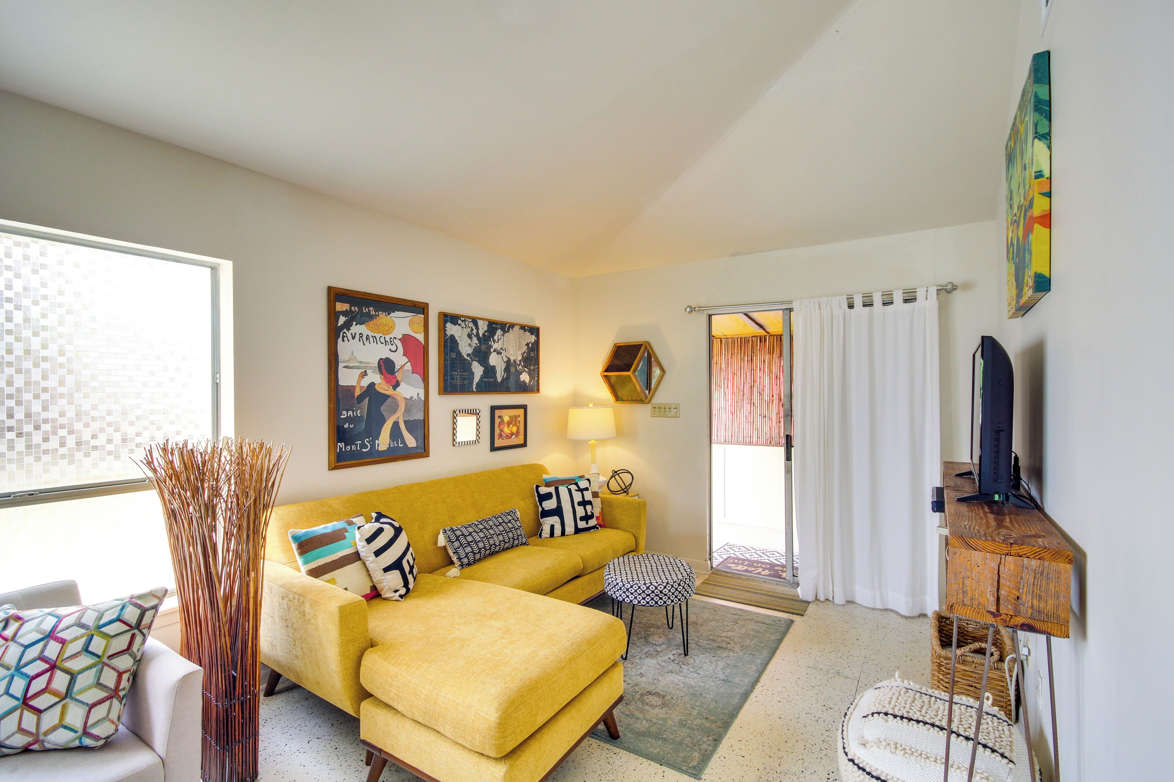 A living room with a yellow sofa, a coffee table, a TV on a wooden stand, and a window with curtains.