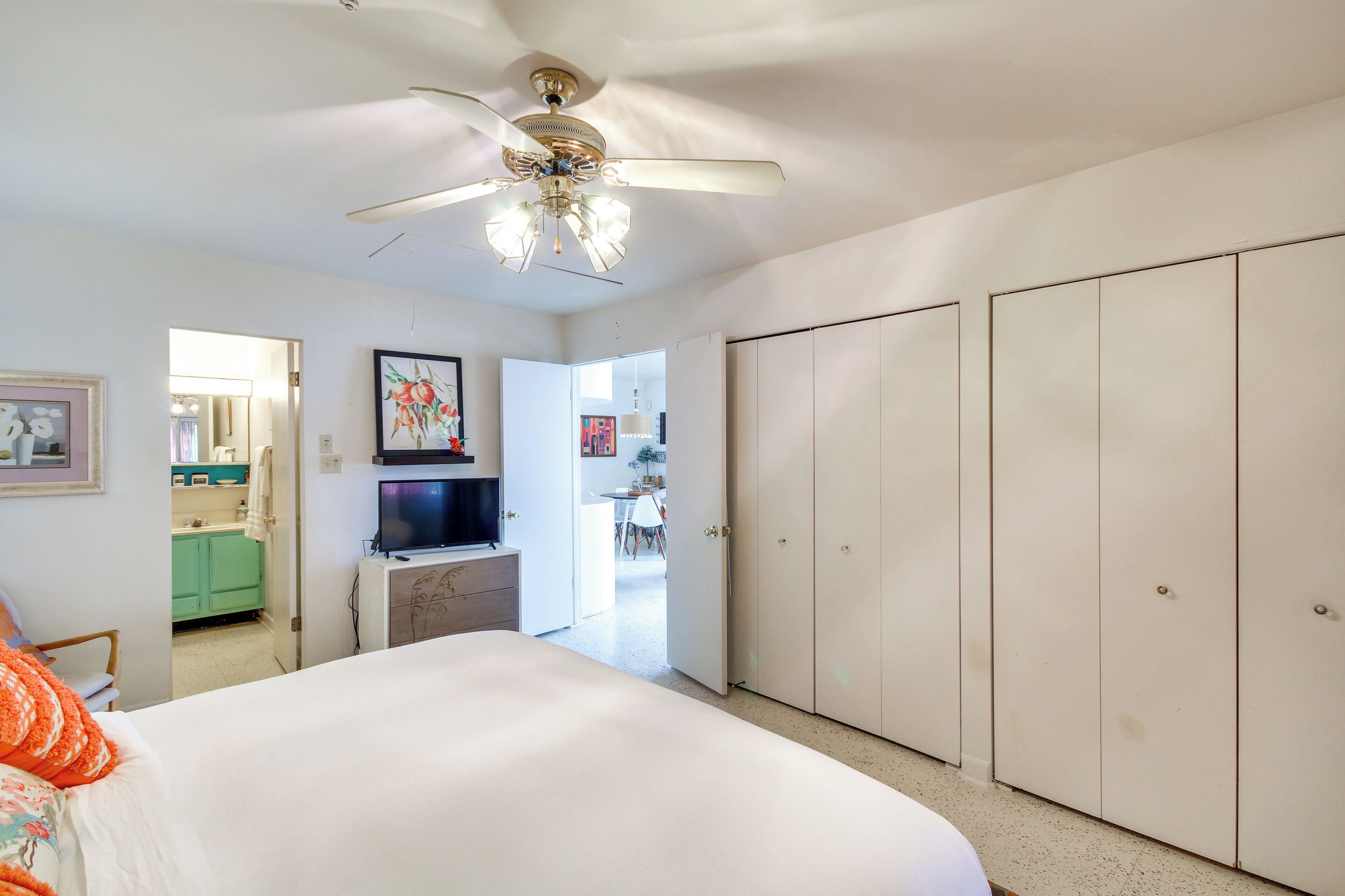 A bedroom with a bed, a ceiling fan, a TV, a wardrobe, and a kitchen area visible through an open door.