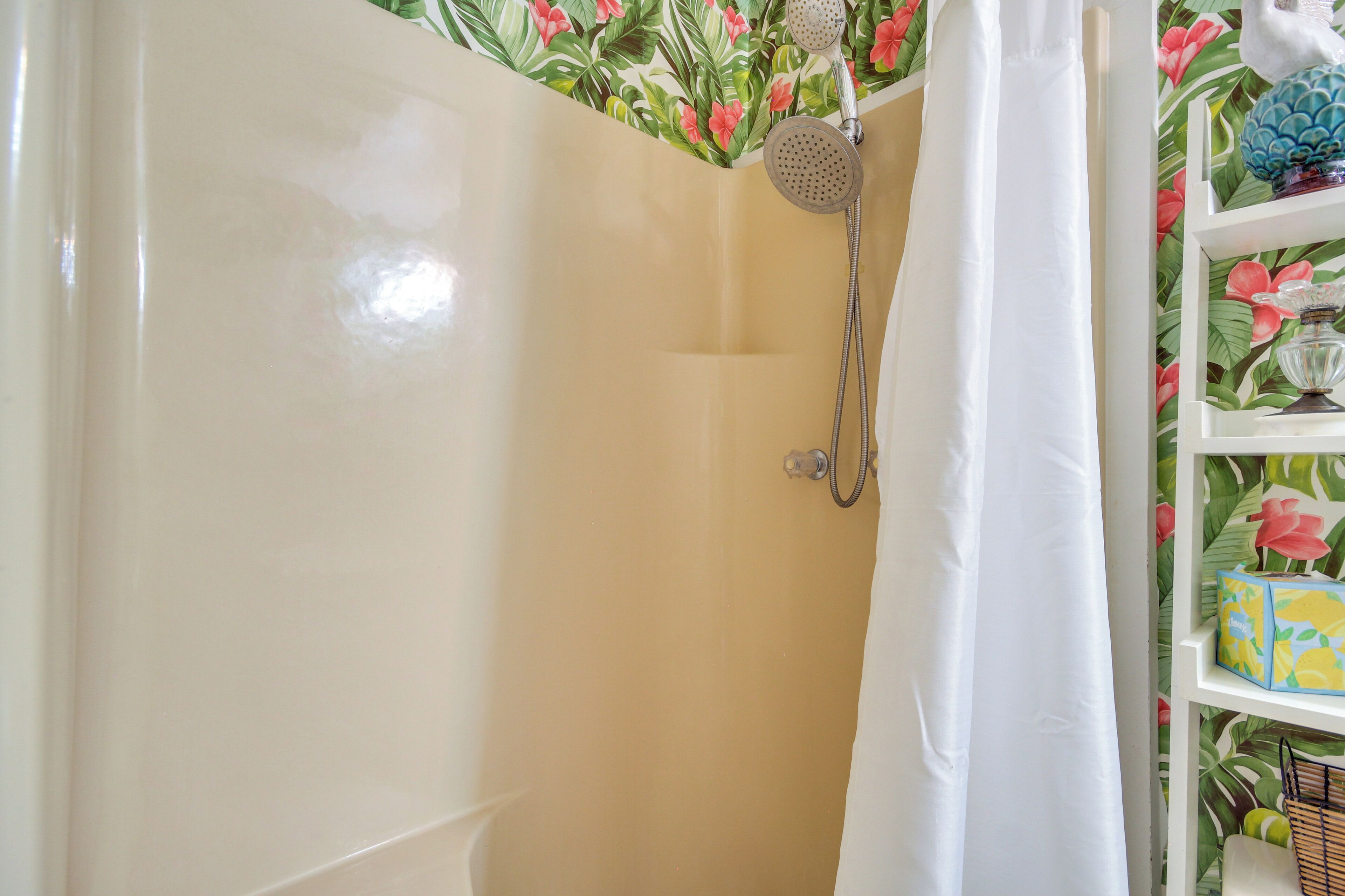 A bathroom with a shower, a white curtain, and a shelf with a floral wallpaper background.