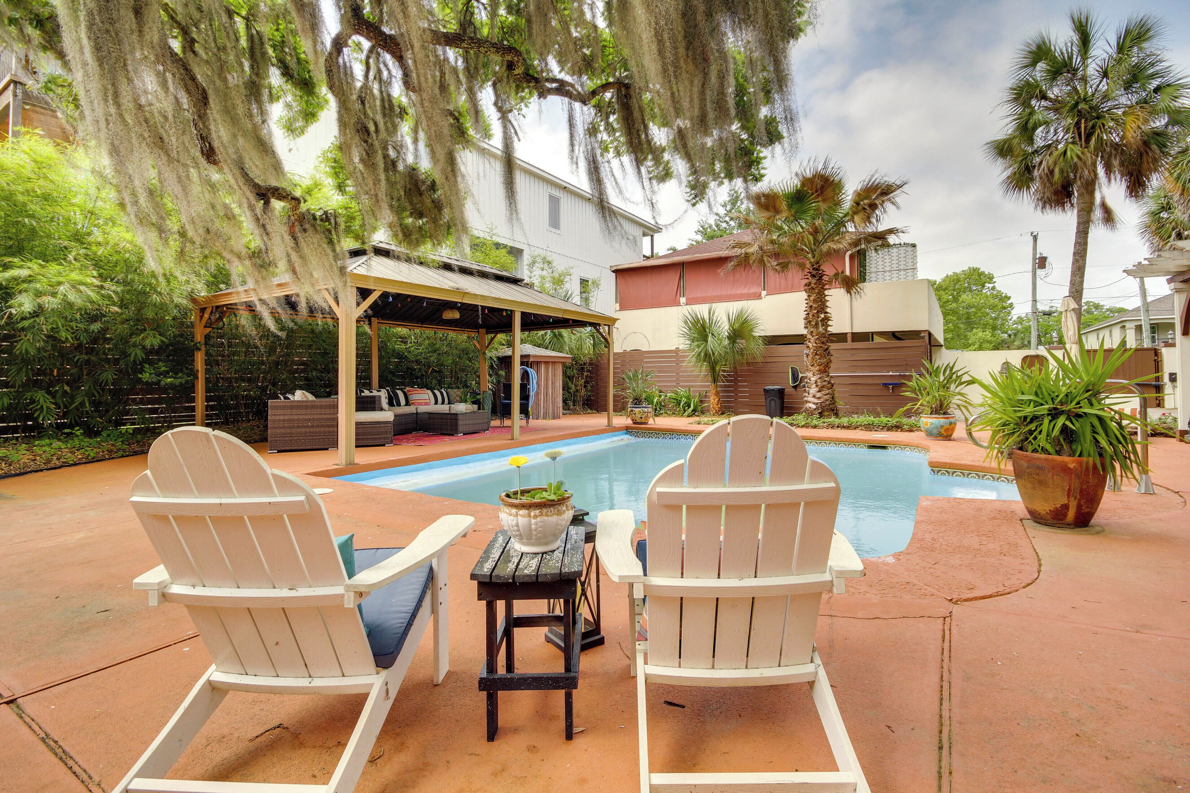 A backyard with a pool, two Adirondack chairs, a covered seating area, and a house in the background.