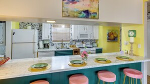 Una cucina con piano di lavoro in marmo, mobili bianchi, dettagli color acquamarina, sgabelli da bar rosa e un quadro astratto dai colori vivaci.