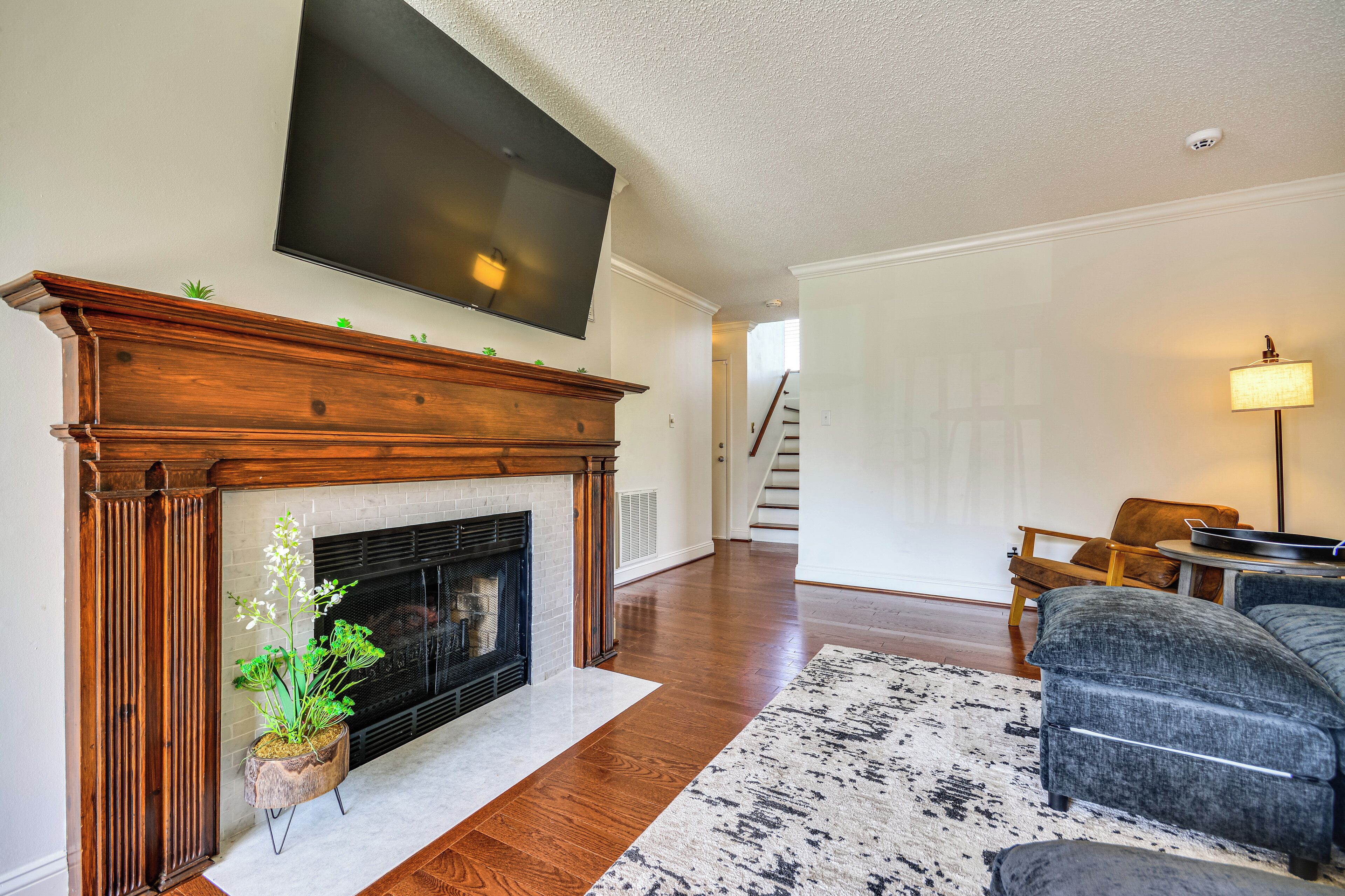 A living room with a fireplace, a flat-screen TV mounted above it, a staircase in the background, and a gray sofa.