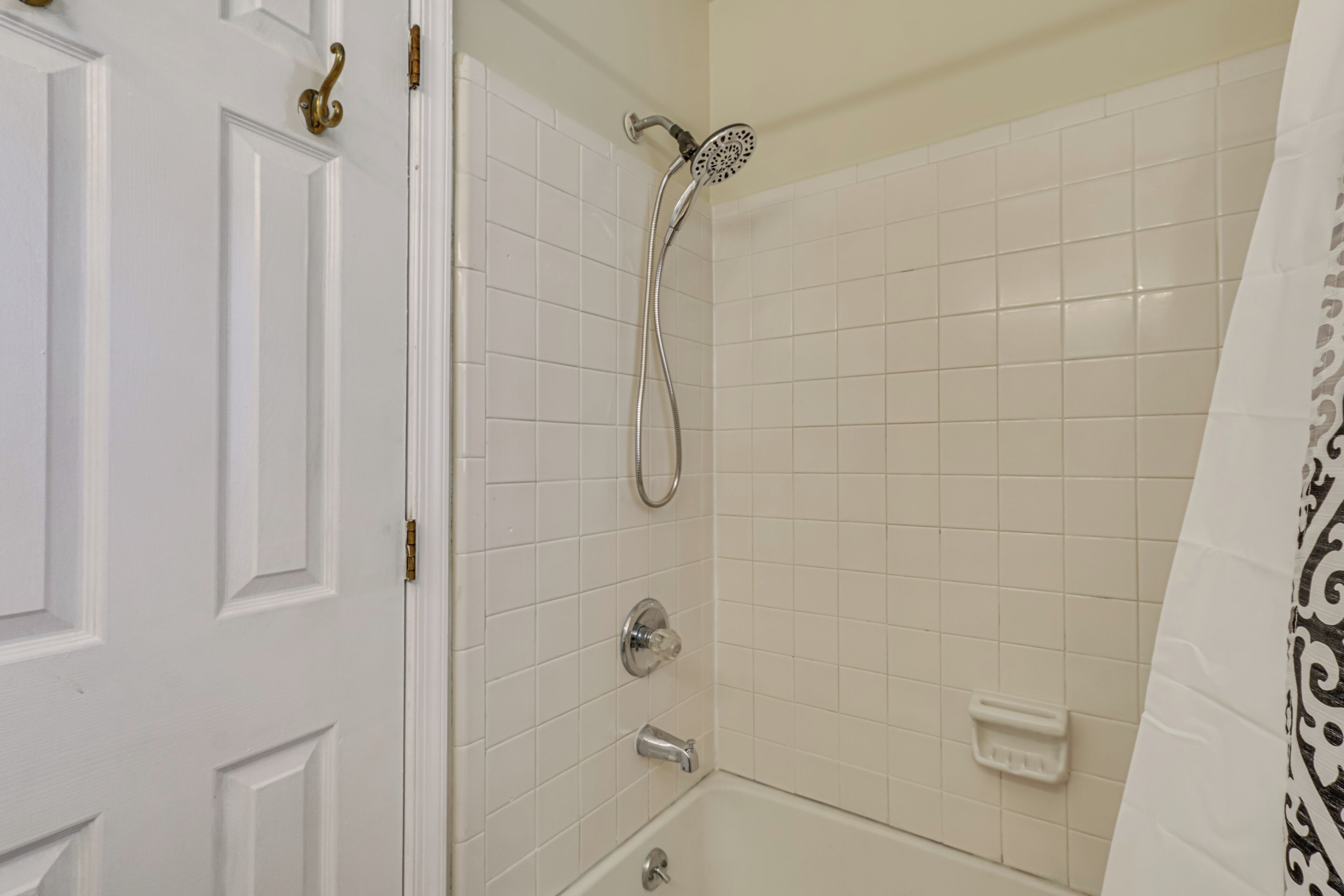 Un baño con paredes de azulejos blancos, ducha con cabezal de ducha manual, inodoro y lavabo con jabonera.