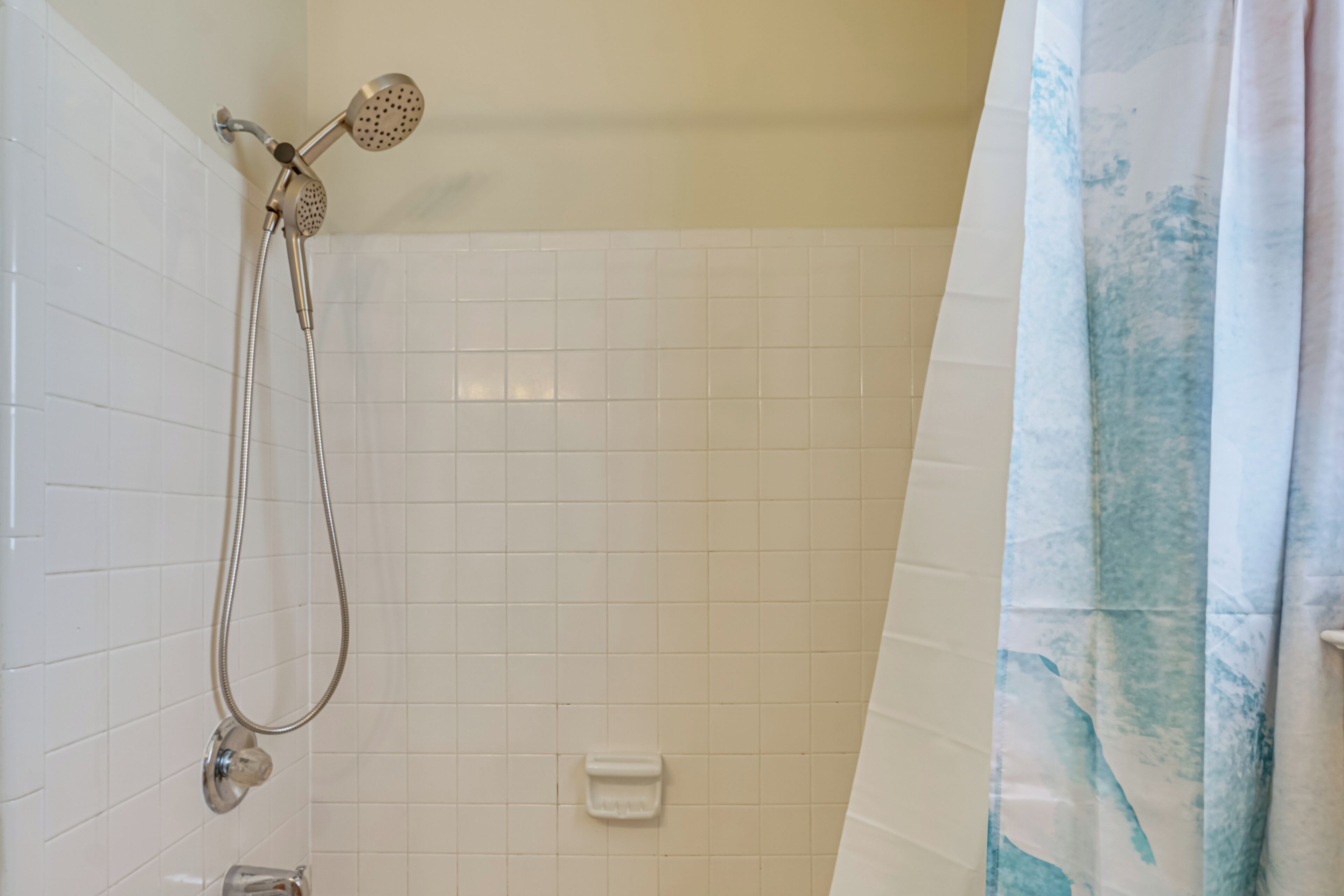 A shower with a handheld showerhead and a handheld hose, a white tiled wall, and a blue and white shower curtain.
