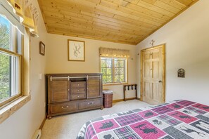 Un dormitorio con techo de madera, una cama grande con colcha estampada, cómoda de madera, ventana con vista y una puerta con una cruz.