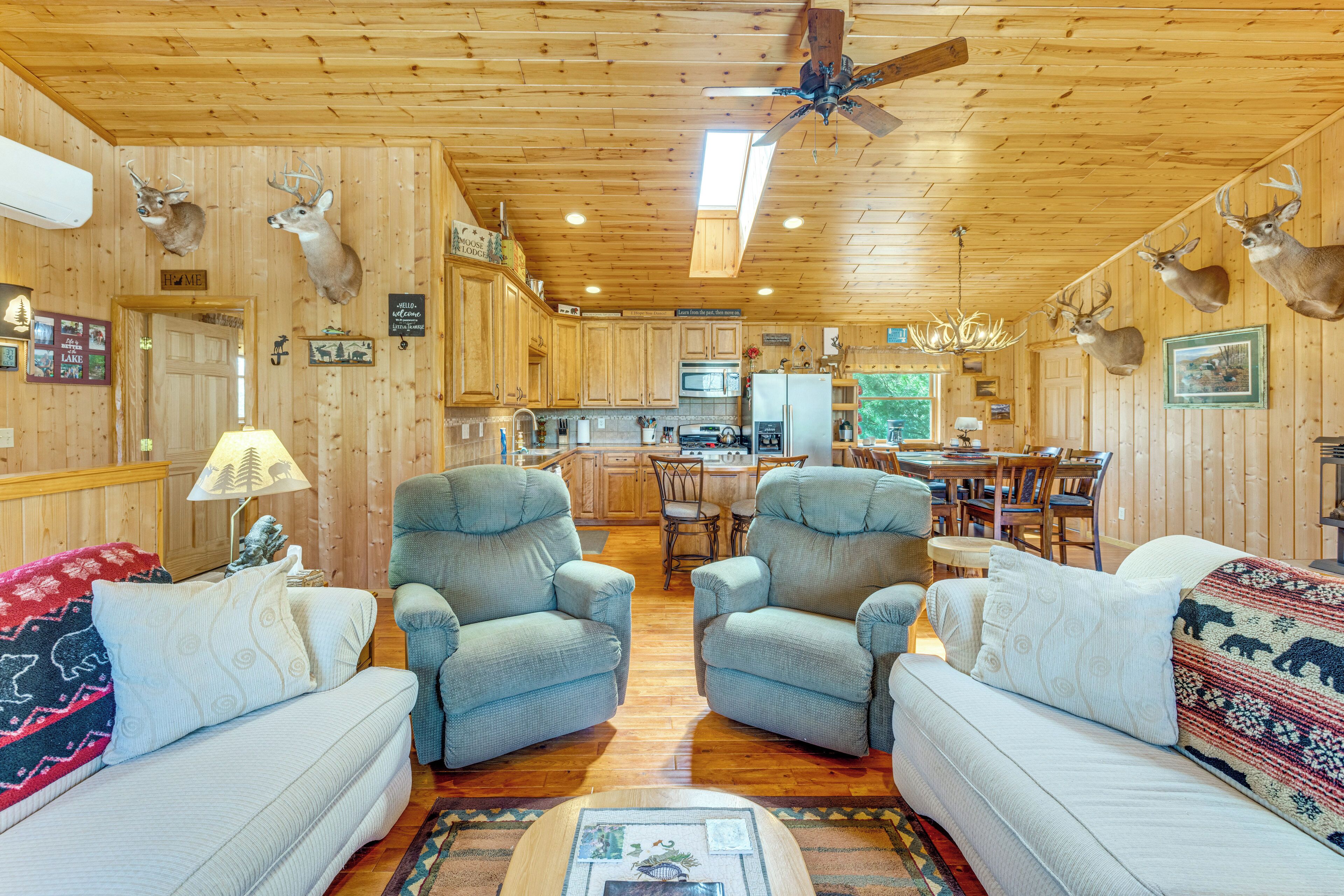 A wooden cabin interior with two sofas, a dining table, chairs, a kitchen area, and mounted animal heads on the walls.